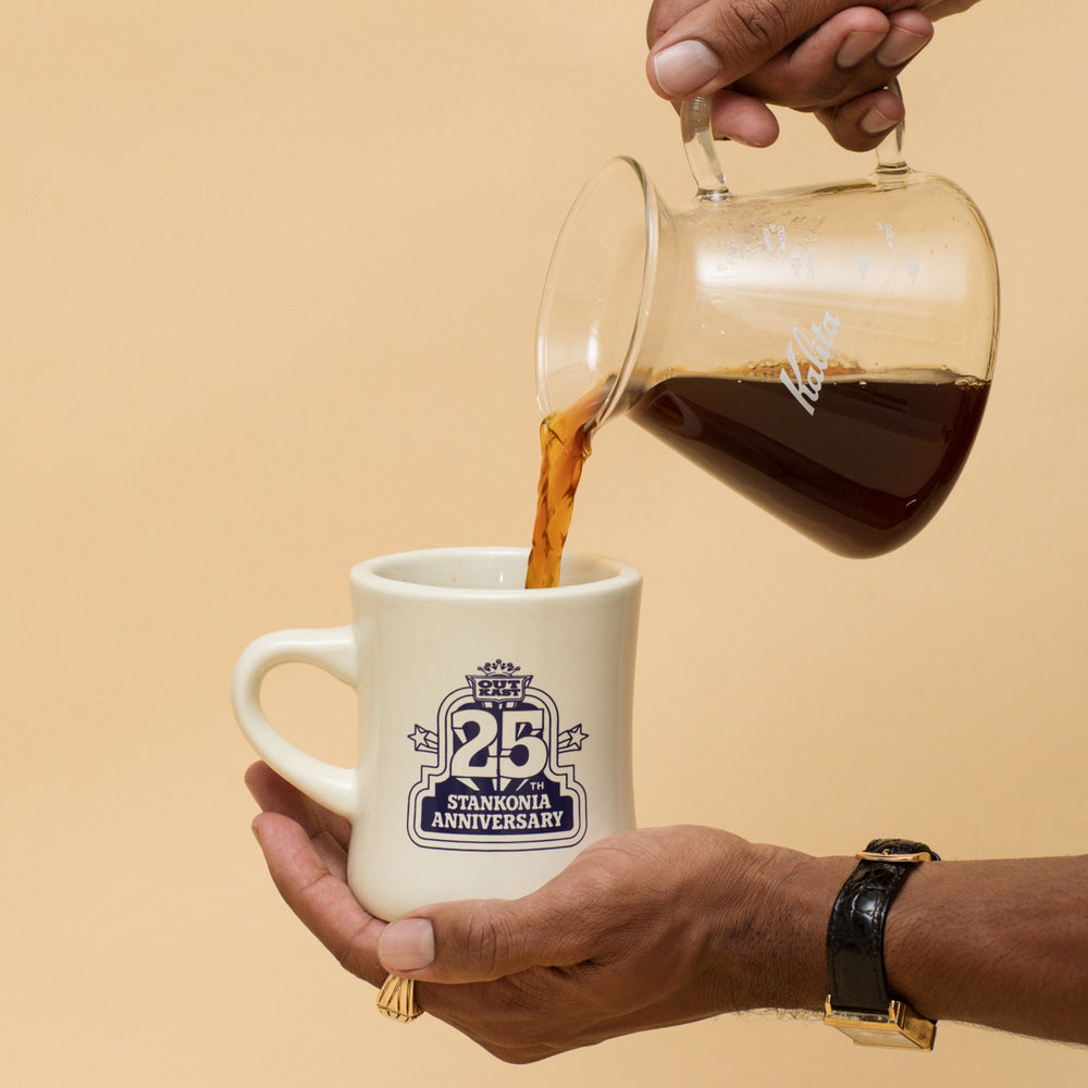
                  
                    Person pouring coffee from a glass carafe into a white mug with 'Stankonia Anniversary' design on a beige background
                  
                