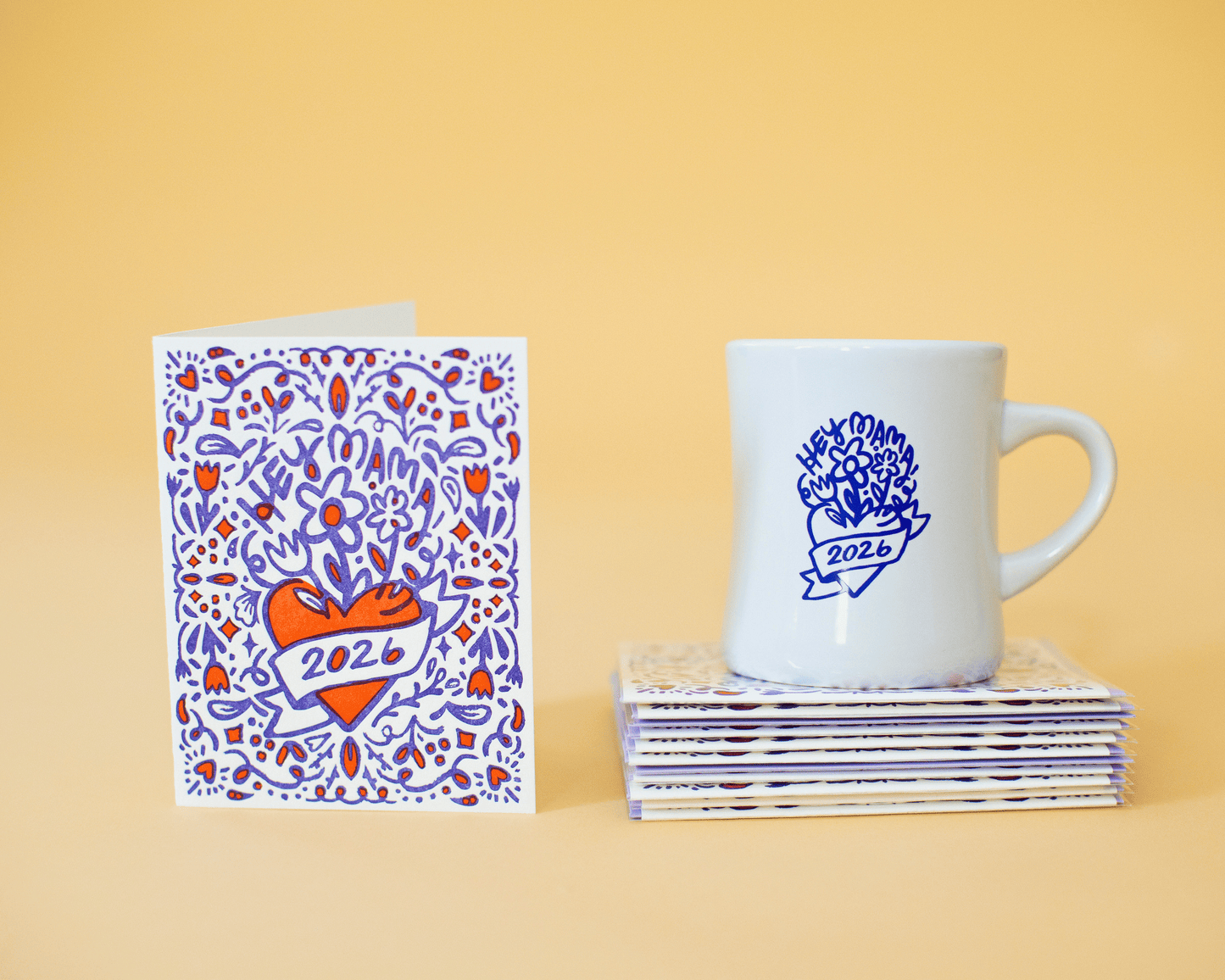 White mug with blue '2026' design and patterned card on a yellow background